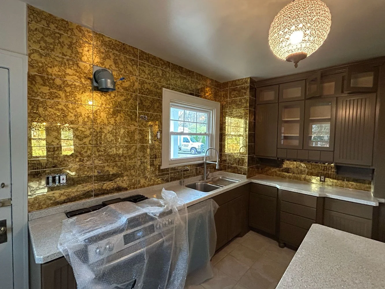 A kitchen with brown cabinets, a window, a tan speckled countertop, and a gold-patterned tiled wall. There is a stove covered in plastic, a double sink, and a ceiling light fixture.