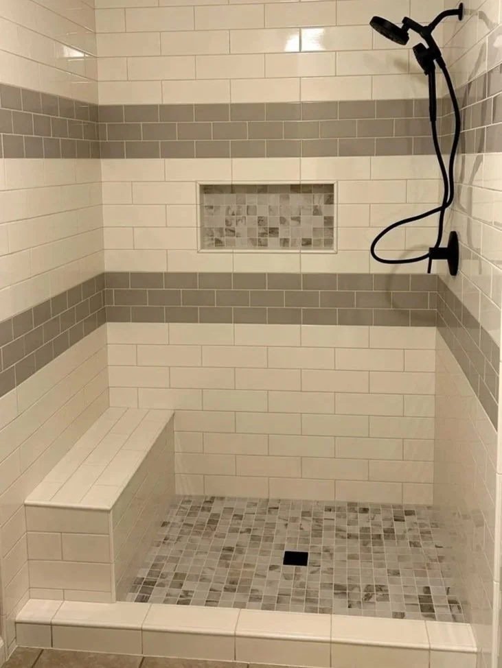 Custom tiled walk-in shower with ivory subway tile walls, gray accent stripe, mosaic tile floor, built-in bench, recessed niche, and matte black shower fixtures.