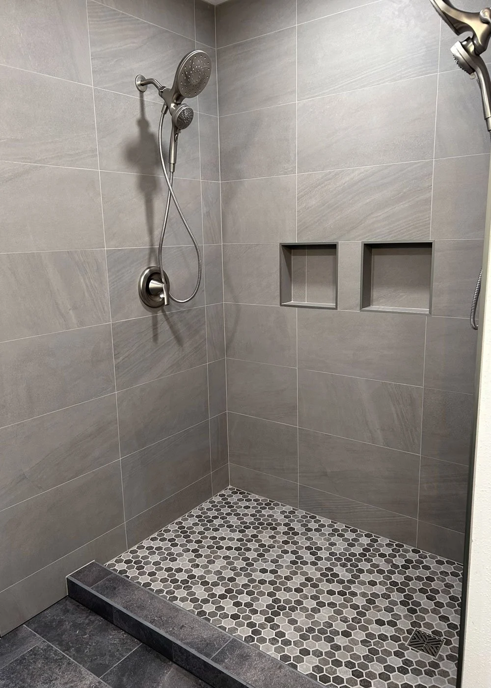 A modern shower stall with gray tiled walls, a handheld showerhead, and two built-in recessed shelves.