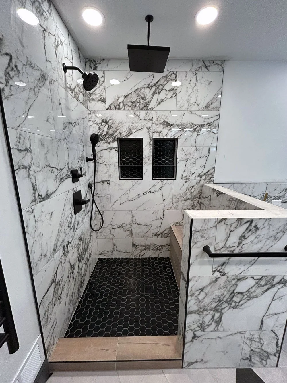 Modern walk-in shower with white marble walls, black shower fixtures, two niche shelves, black hexagonal floor tiles, and a rainfall showerhead.