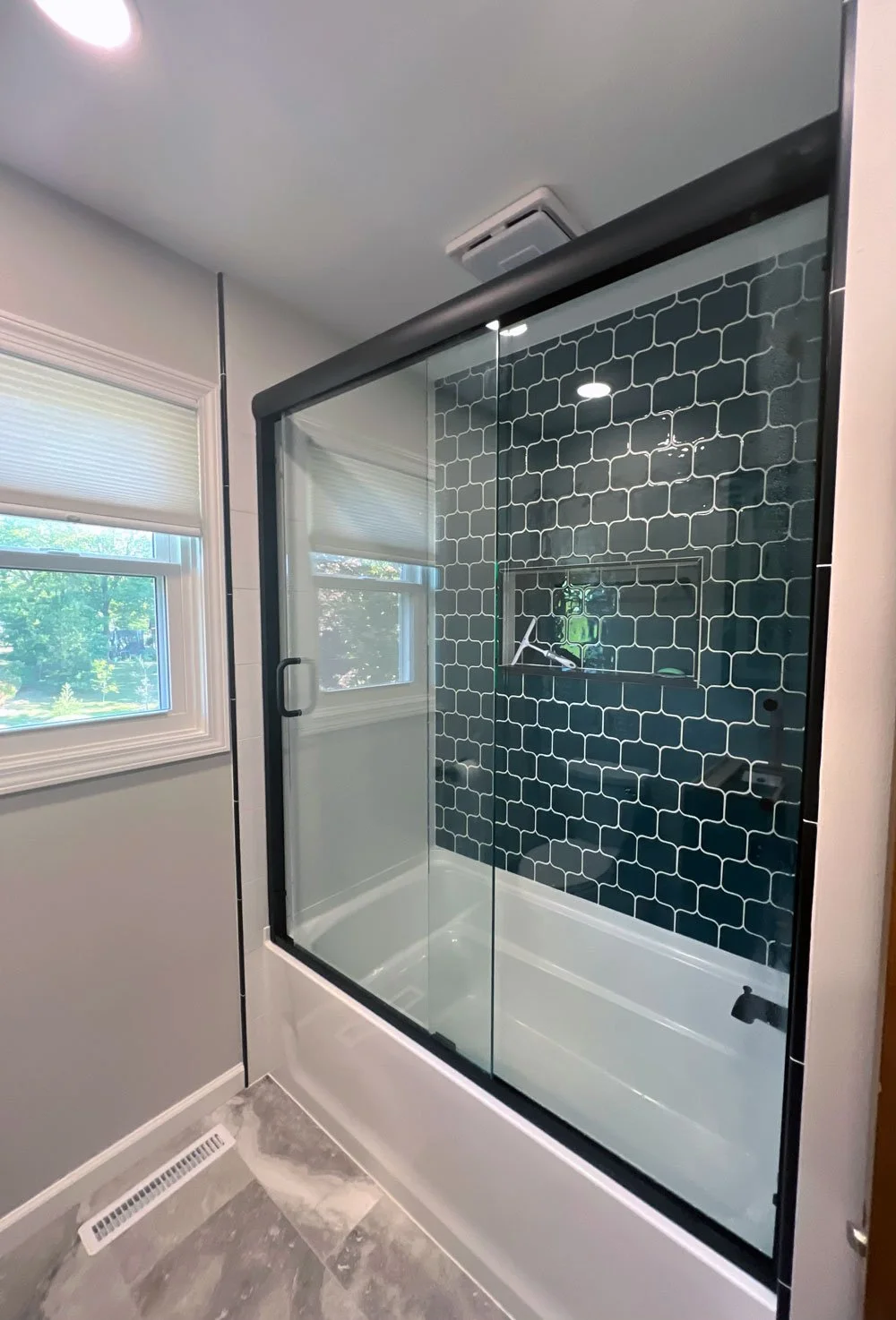 Glass shower with black tile backsplash, a built-in niche, in a bathroom with two windows and a marble floor.