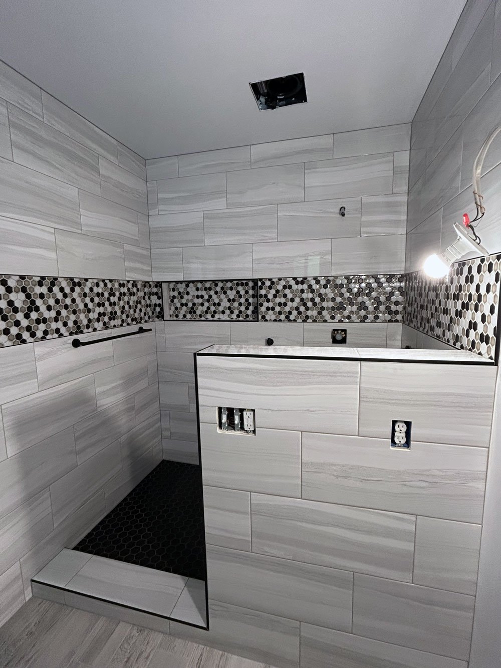 Bathroom under renovation with tiled walls in gray and white tones, a built-in shower area with black hexagonal floor tiles, electrical outlets, and an overhead light fixture.