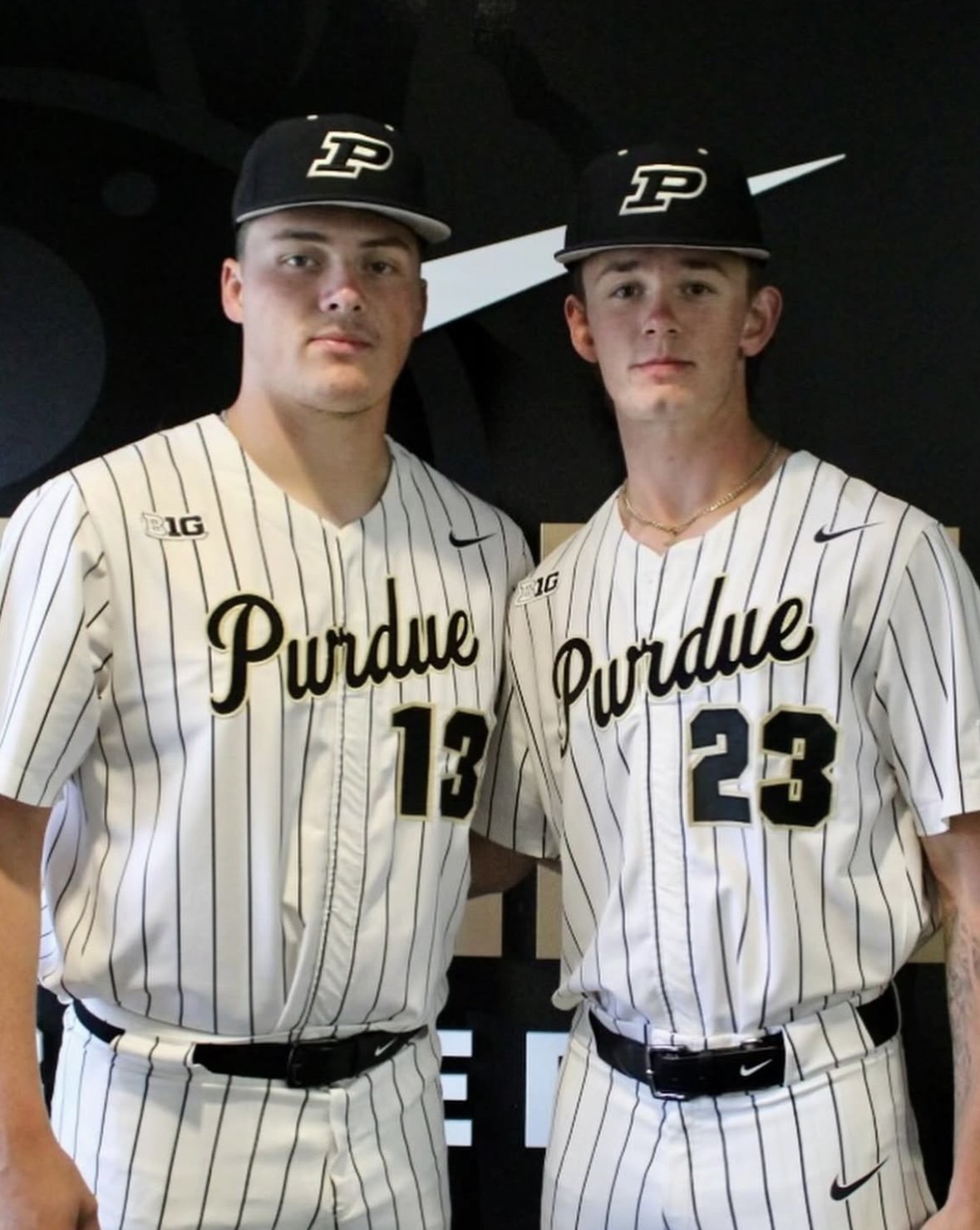 Two young men in Purdue baseball uniforms standing together, both wearing black caps with the Purdue logo, and white pinstripe jerseys with 'Purdue' written across the chest and numbers 13 and 23.