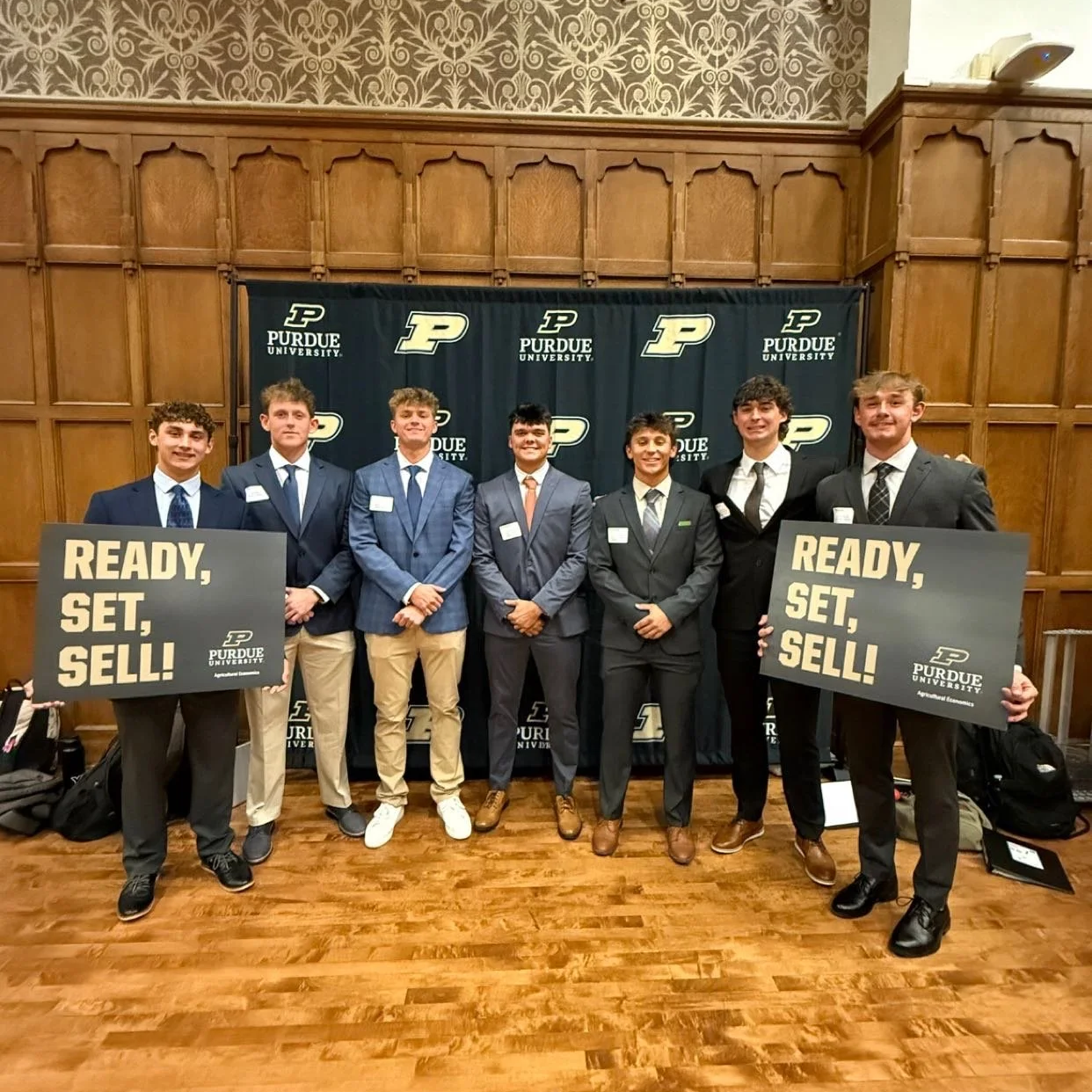 Seven young men dressed in suits standing in a line in front of a Purdue University backdrop, holding signs that say 'READY, SET, SELL!' at an indoor event.