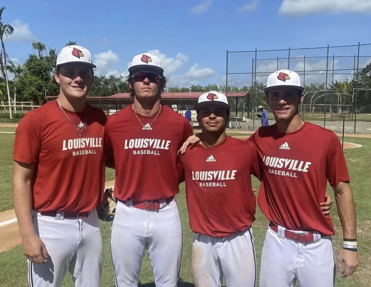 Four baseball players wearing red shirts with 'Louisville Baseball' and white pants, standing together on a baseball field under a partly cloudy sky, posing for a team photo.