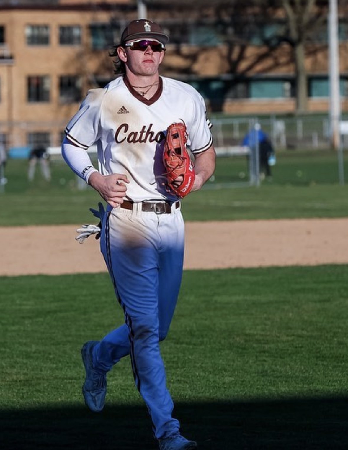 A young male baseball player is walking on a baseball field. He is wearing a white and blue uniform with the word 'Catholic' on the front, a brown cap with a logo, sunglasses, and a red glove.