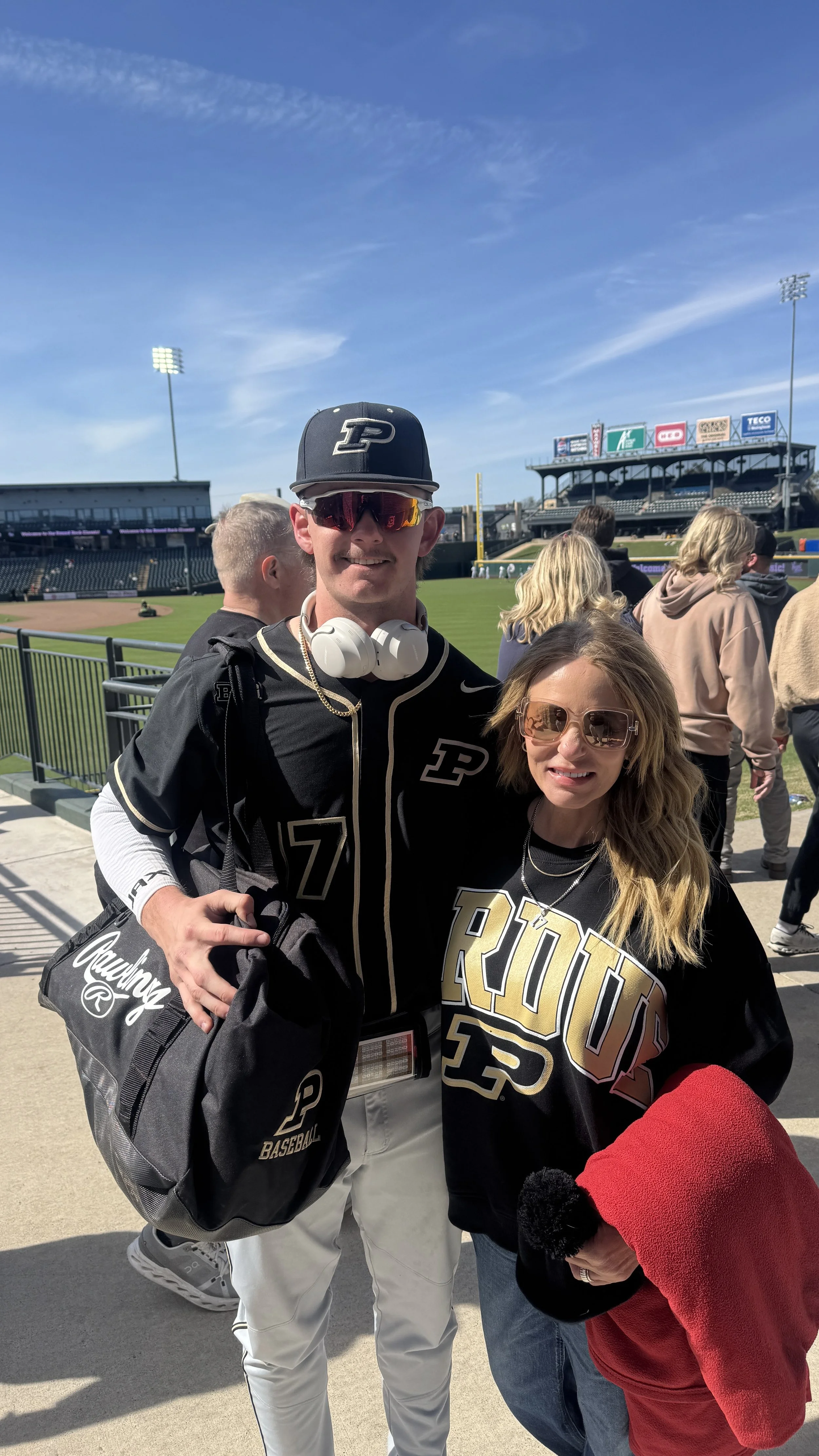 Two people, a man and a woman, standing outdoors near a baseball field, wearing Purdue University sports apparel. The man is holding a bag with Purdue branding, and the woman is holding a red jacket. The background shows fans and the stadium with bri