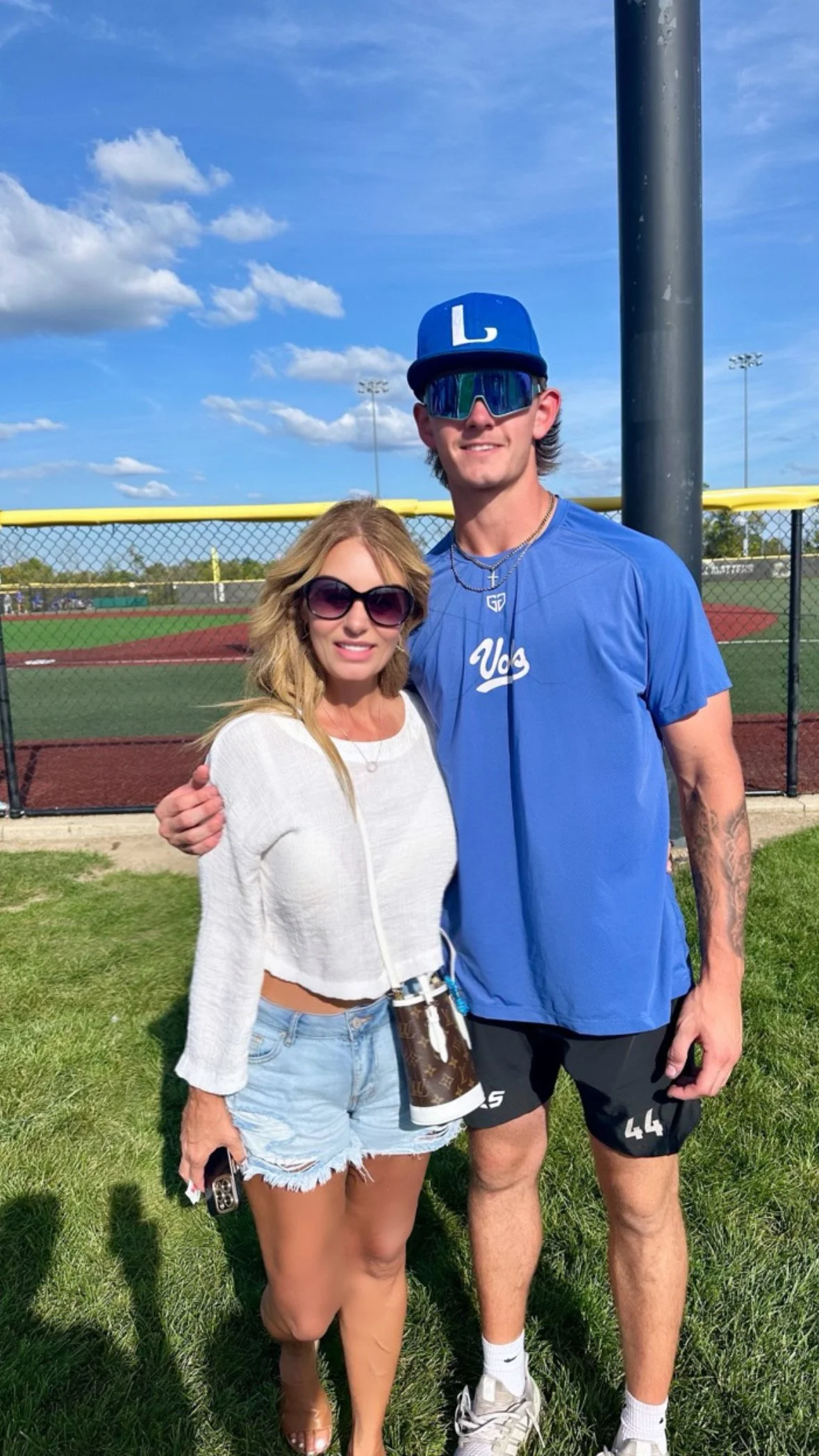 A woman and a tall man standing together on grass near a baseball field, with a fence and blue sky in the background. The woman is wearing sunglasses, a white crop top, and denim shorts. The man is wearing sunglasses, a blue cap with an 'L' on it, a 