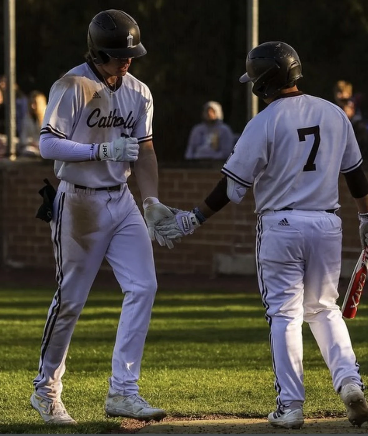 Two baseball players in white uniforms and black helmets shake hands on a baseball field during a game, with spectators in the background.