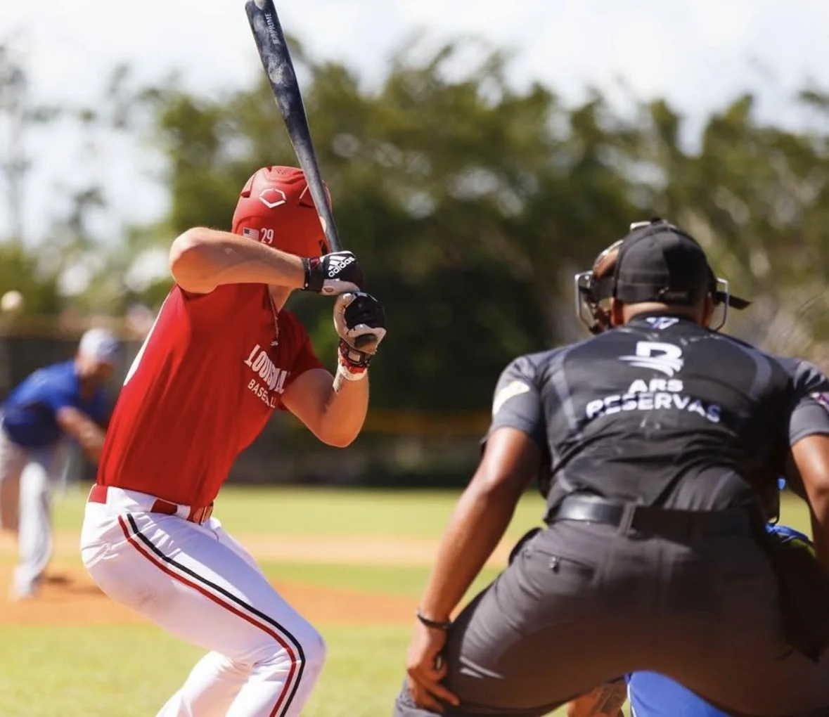 A baseball batter in a red uniform and helmet prepares to hit a pitch during a game, with an umpire in black crouching behind home plate.