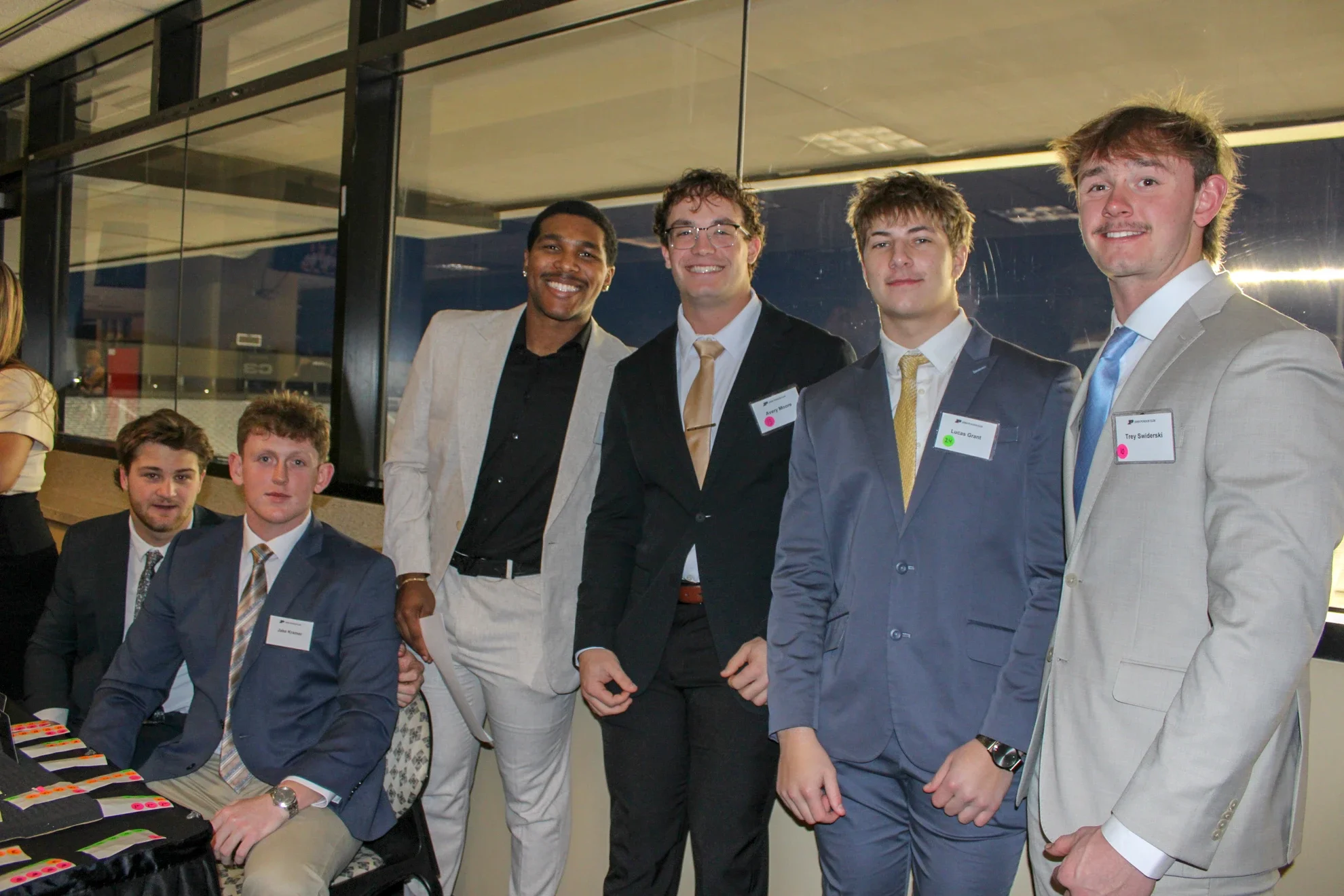 Group of six young men dressed in business suits at a professional event, some standing and some seated, with name tags and a table with colorful items in front of them.