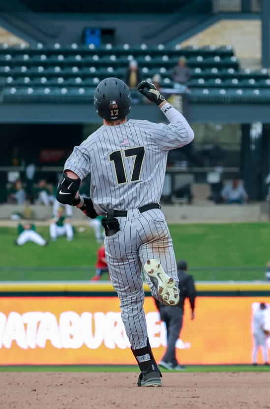 Baseball player wearing gray pinstripe uniform with the number 17, running on a baseball field with a black helmet, and a green outfield with a digital scoreboard in the background.