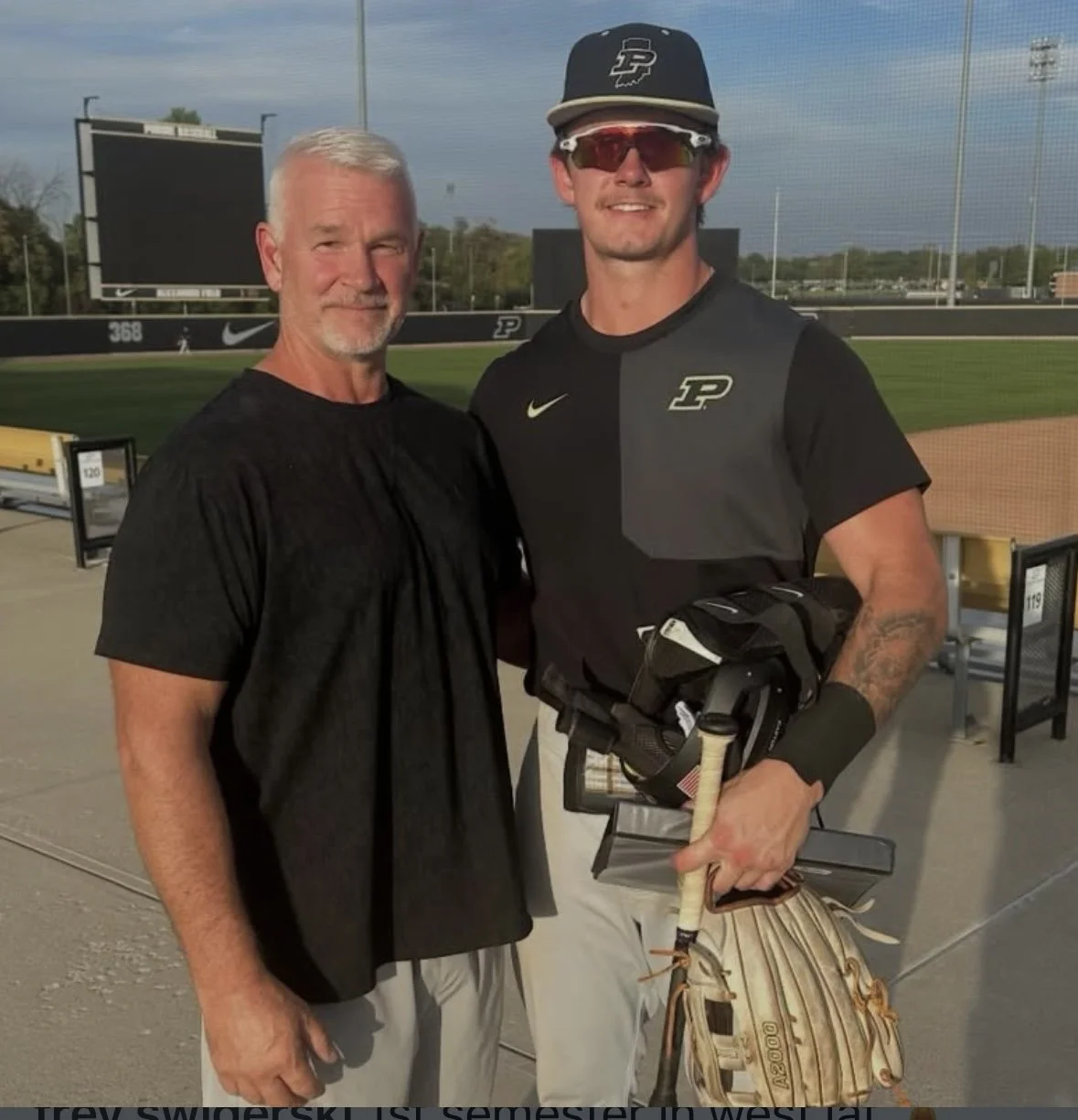 A man in a black sports shirt with the Purdue University logo, wearing sunglasses and a cap, holding a baseball glove and bat, standing with an older man in a black shirt at a baseball stadium.