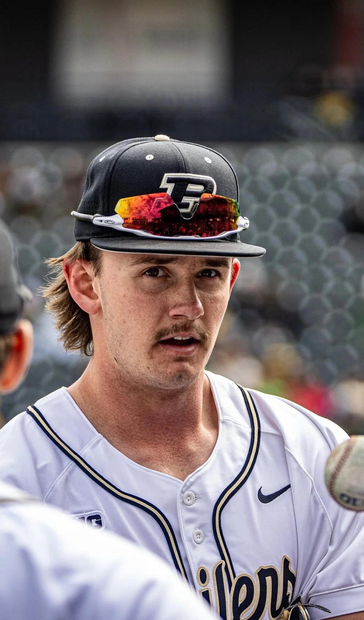 Close-up of a baseball player with a mustache wearing a black cap with a logo, sunglasses on top, and a white jersey with black and gold accents, during a game or practice.