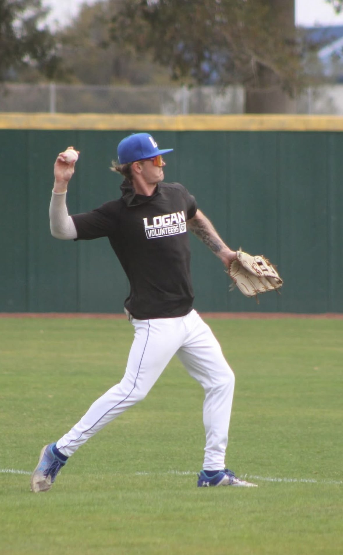 A baseball player in a black shirt, white pants, and a blue cap throws a baseball on a field.