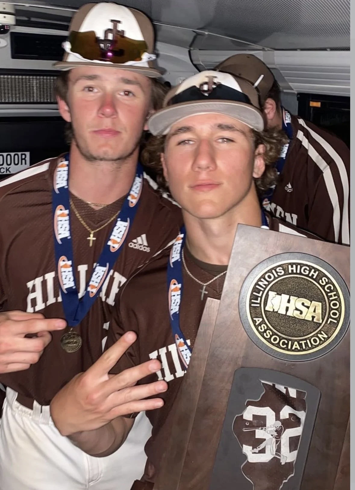 Two young men in brown football jerseys with 'Illinois' written on them, wearing medals and baseball caps, holding a wooden plaque with the Illinois High School Association logo and Michigan State-shaped design, celebrating their achievement.
