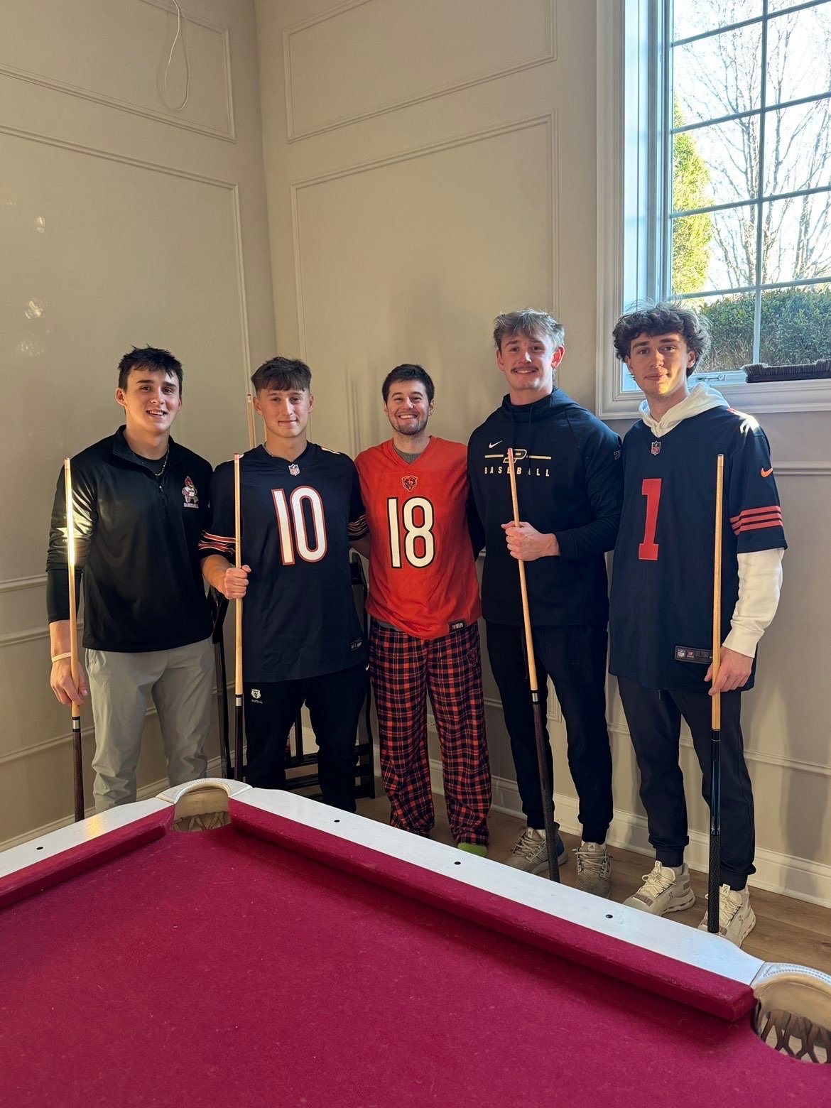 Five young men standing indoors next to a pool table, holding pool cues, smiling, with some wearing sports jerseys and casual clothing, near a window showing trees outside.