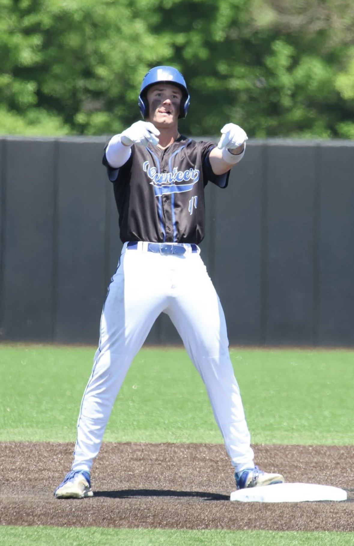A baseball player standing on bases during a game, dressed in a black uniform and helmet, with his hands raised as if signaling or celebrating.