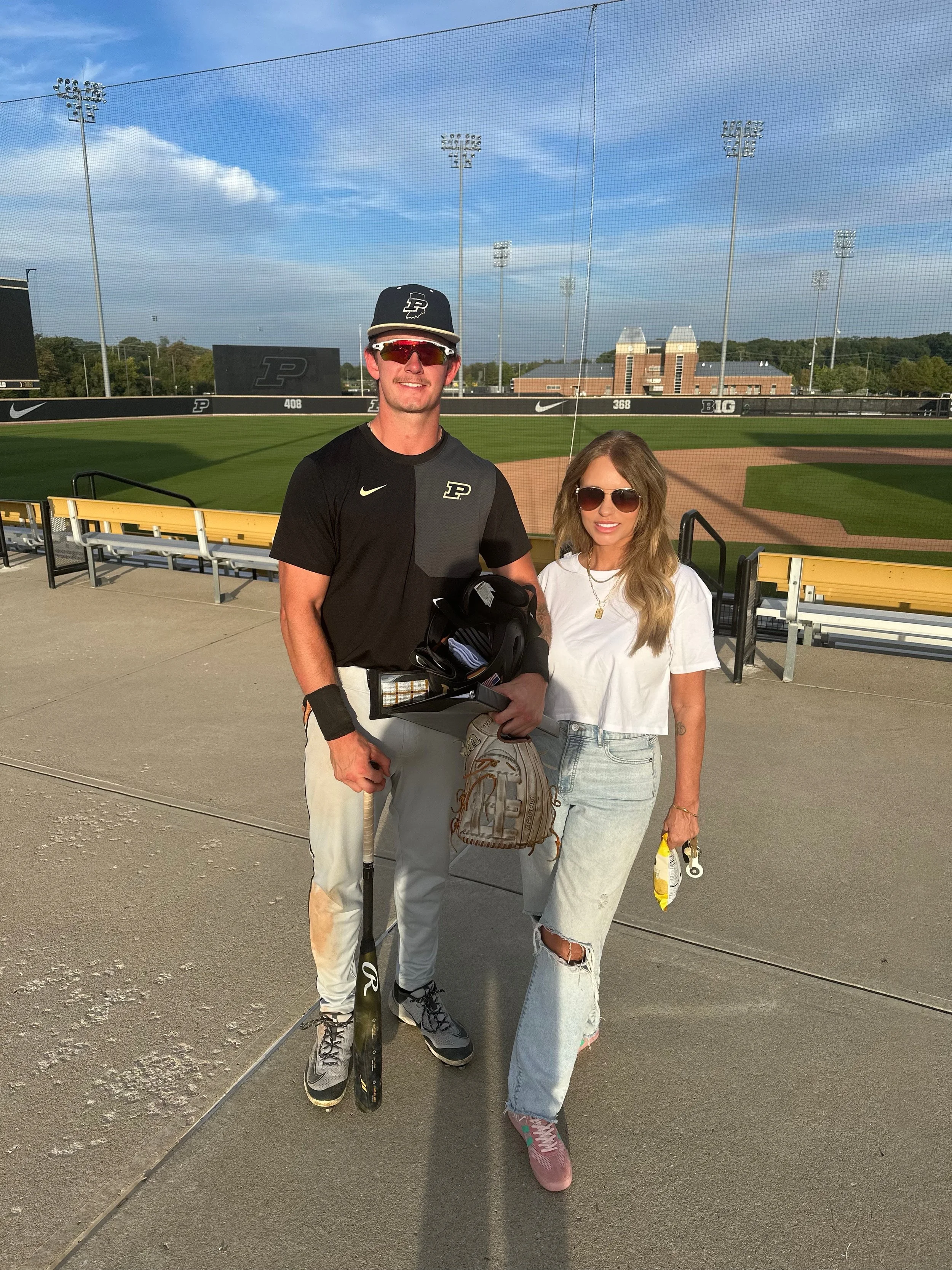 A man and woman standing on a baseball field's dugout area, with the baseball field and stadium in the background. The man is wearing a baseball uniform with a cap, sunglasses, and holding a baseball bat. The woman is wearing casual clothing, includi