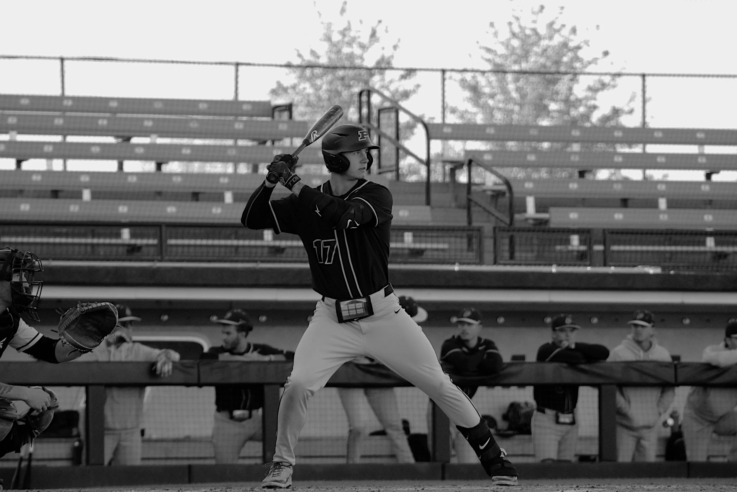 A baseball player stands at home plate, preparing to swing at a pitch during a game. The player is wearing a helmet and uniform with the number 17, holding a bat in a ready position. There are spectators or team members watching from the dugout in th