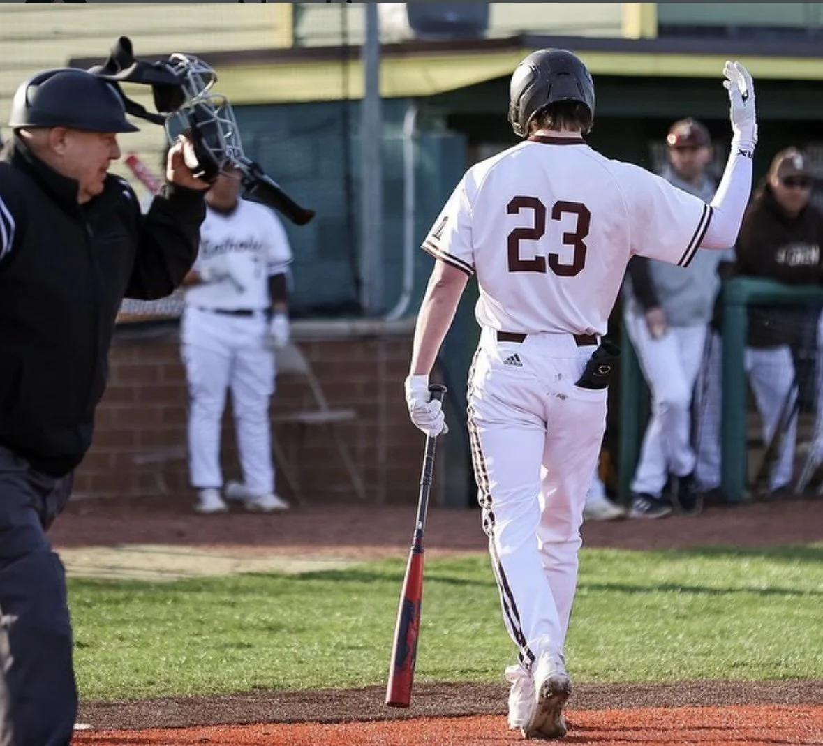 A baseball player wearing a white uniform with the number 23 on the back, holding a bat, walking away from home plate, waving to the crowd after scoring a run during a game, with an umpire and other players in the background.