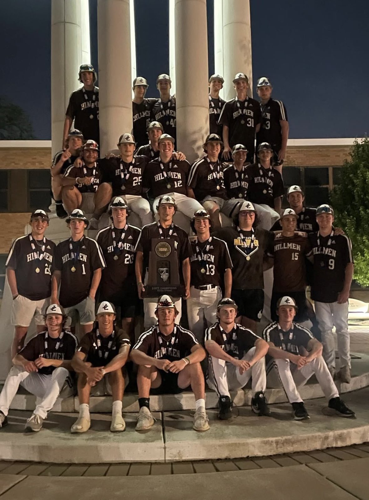 A baseball team named the Hillmen celebrating at night, posing with a championship trophy in front of a monument with tall columns.