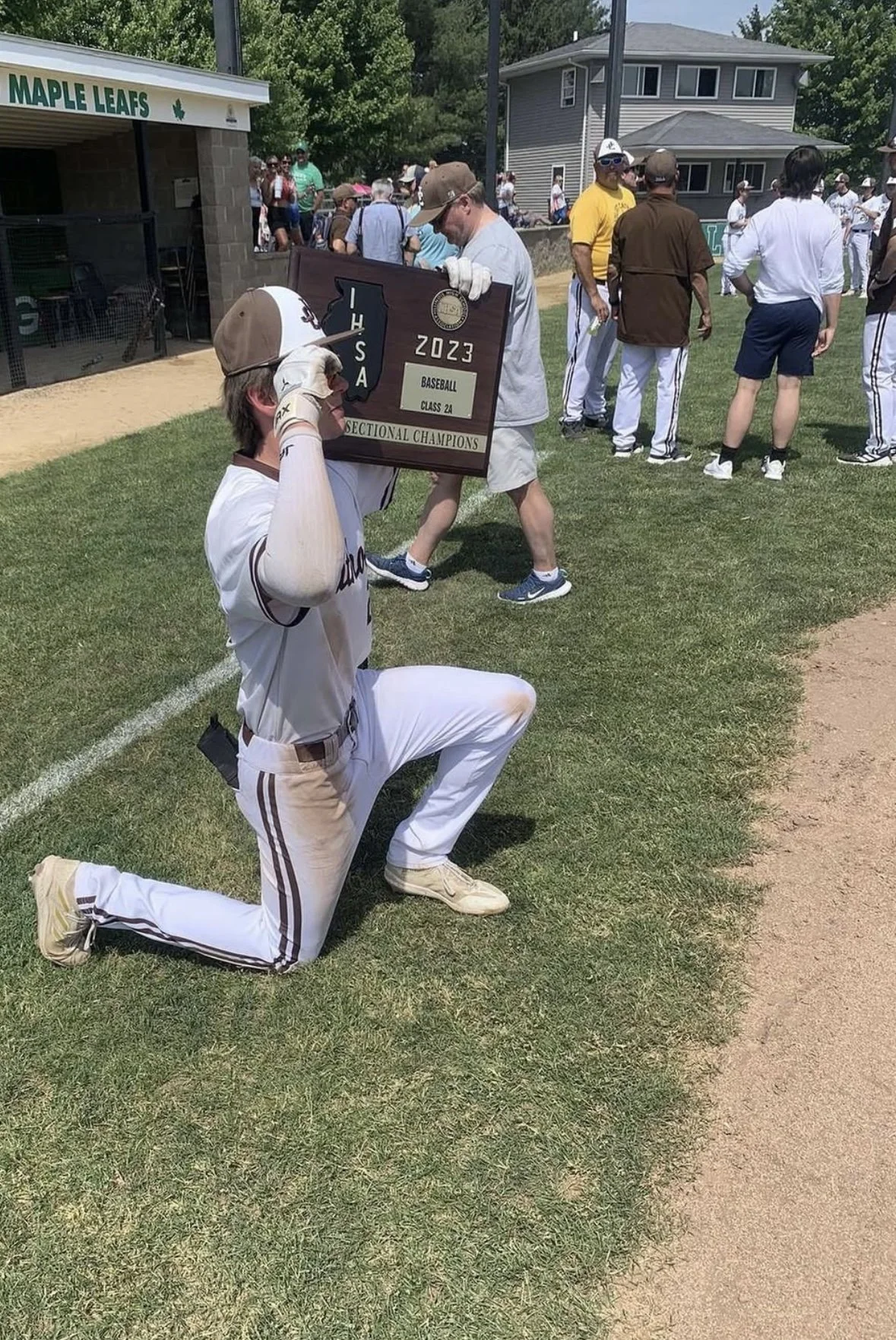 A baseball player kneeling on one knee on the field, holding a championship plaque for the 2023 ISA baseball class 24 sectional champions, with spectators and team members in the background.