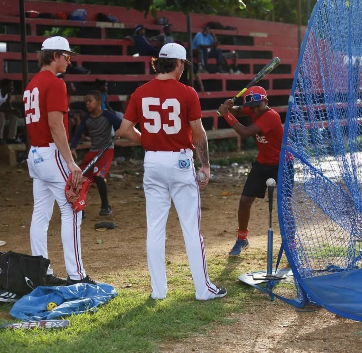 Youth baseball players practicing hitting in a batting cage during daylight. Two players in red jerseys and white pants standing outside the cage, with one holding a bat and the other holding a glove. A boy in the cage wearing cycling goggles is prep