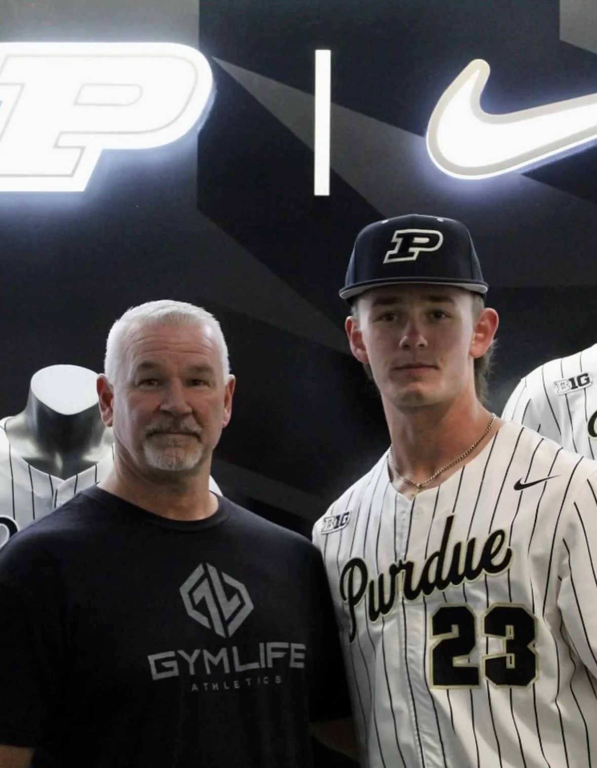 A man and a young man wearing a Purdue baseball uniform and cap, standing in front of a black wall with illuminated Nike and Purdue logos.