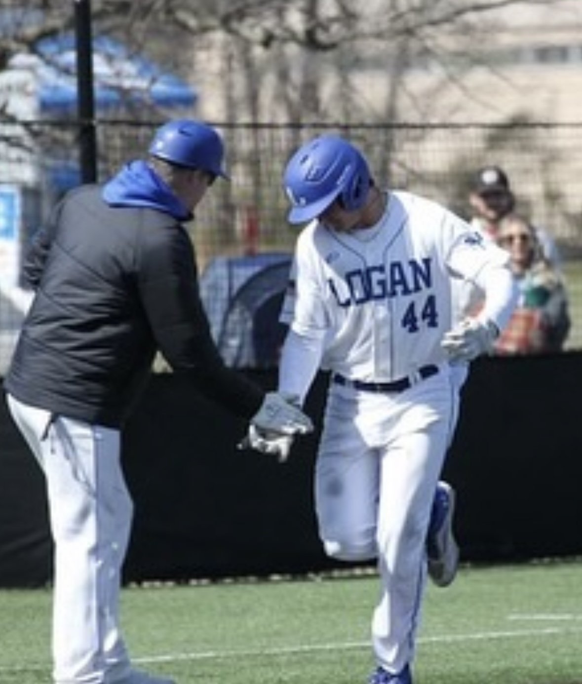 A baseball player in a white uniform with blue lettering and the number 44 is running towards home plate on a baseball field while an umpire in a black jacket and blue helmet is holding a baseball and extending his hand for a handshake or high-five.