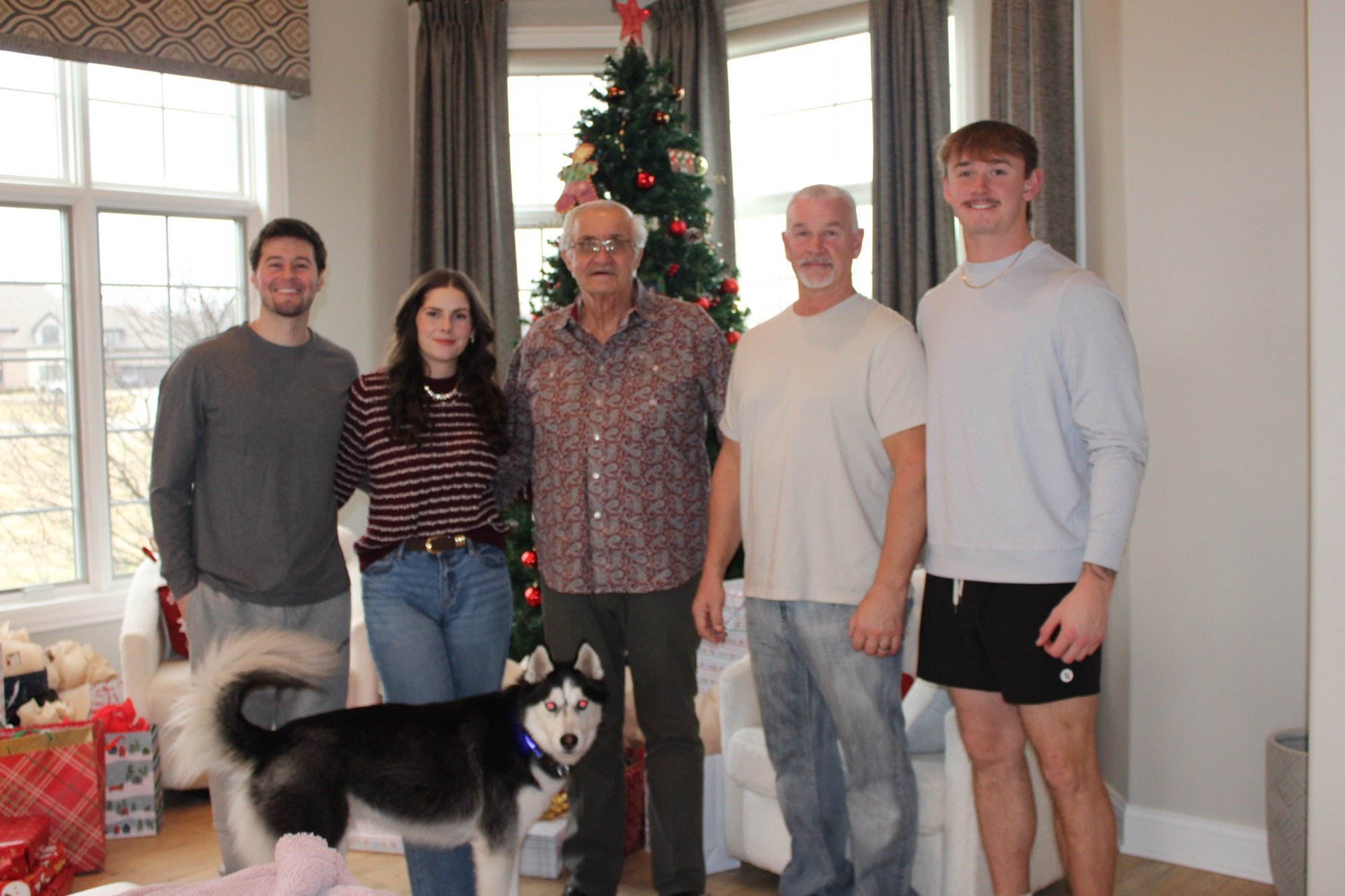 Family of five standing in front of a decorated Christmas tree inside a living room, with a husky dog in the foreground and wrapped presents on the floor.