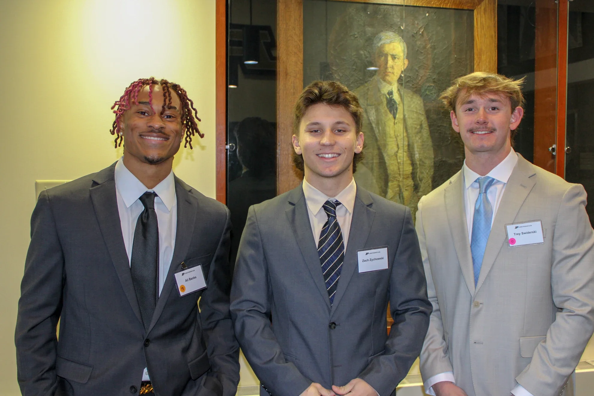 Three young men wearing suits and name tags standing together in front of a framed picture of a man wearing a suit.