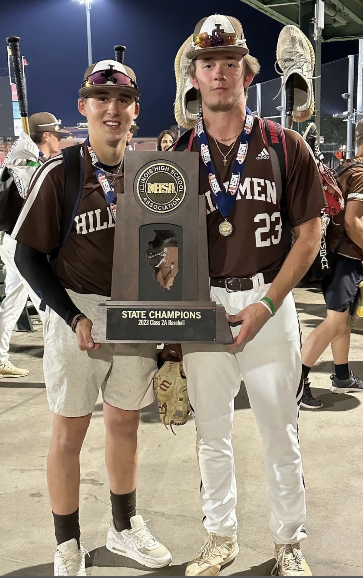 Two young men in sports uniforms holding a trophy that reads 'State Champions 2023 Class 2A Baseball,' celebrating their victory at a high school baseball tournament.