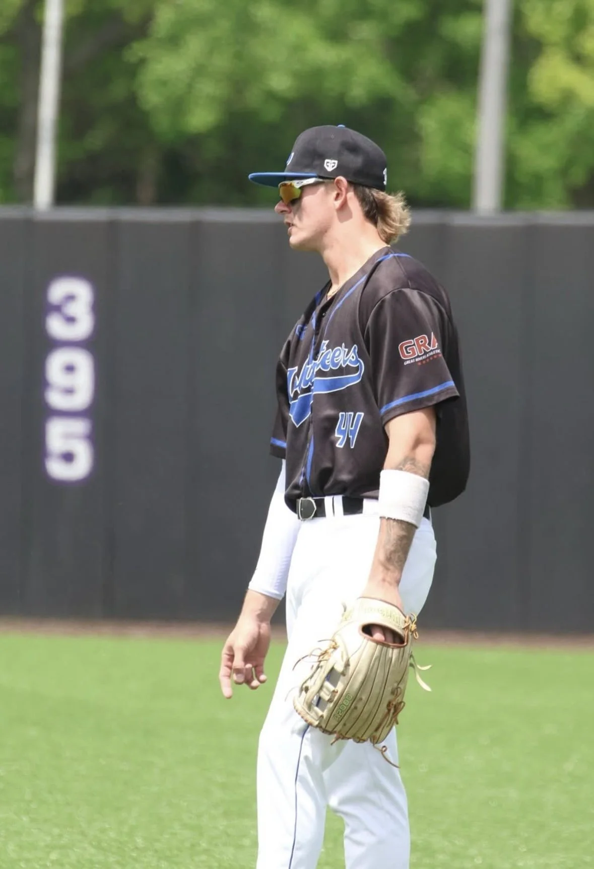 A baseball player standing on the field in a black jersey and white pants, wearing a cap and sunglasses, holding a baseball glove in one hand.
