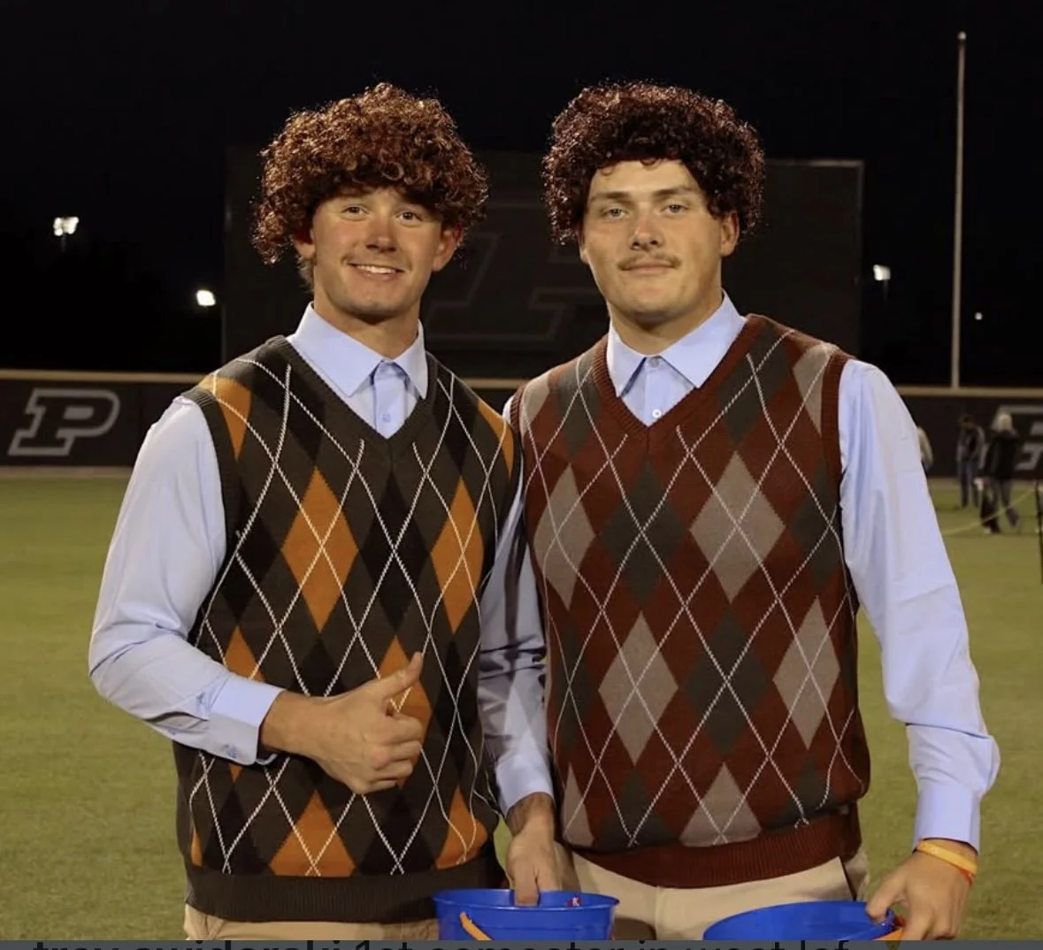 Two young men dressed in collared shirts and argyle vests, standing on a football field at night, smiling and posing for the photo.