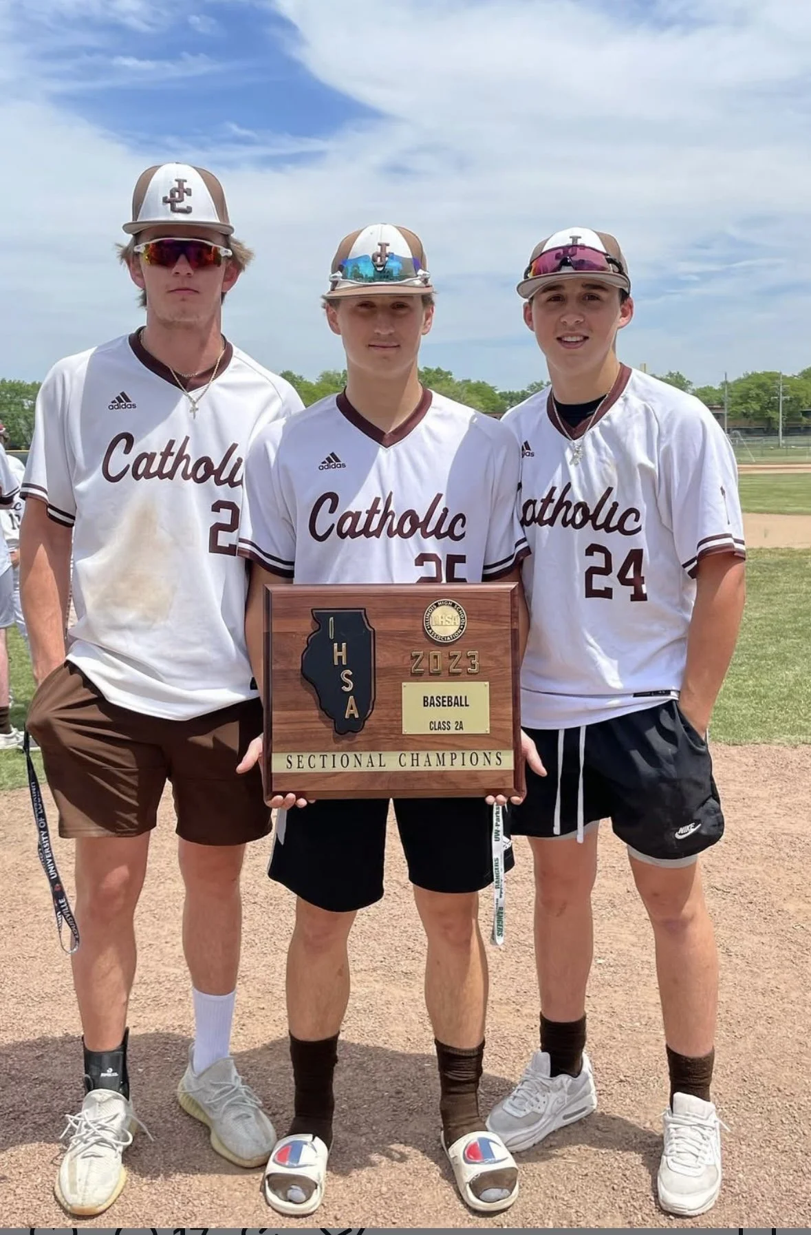 Three teenage boys in baseball uniforms holding a plaque, standing on a baseball field. The plaque reads 'IHSAA 2023 Baseball Class 2A Sectional Champions.' They are wearing white jerseys with 'Catholic' written on them, and their caps have a logo. T