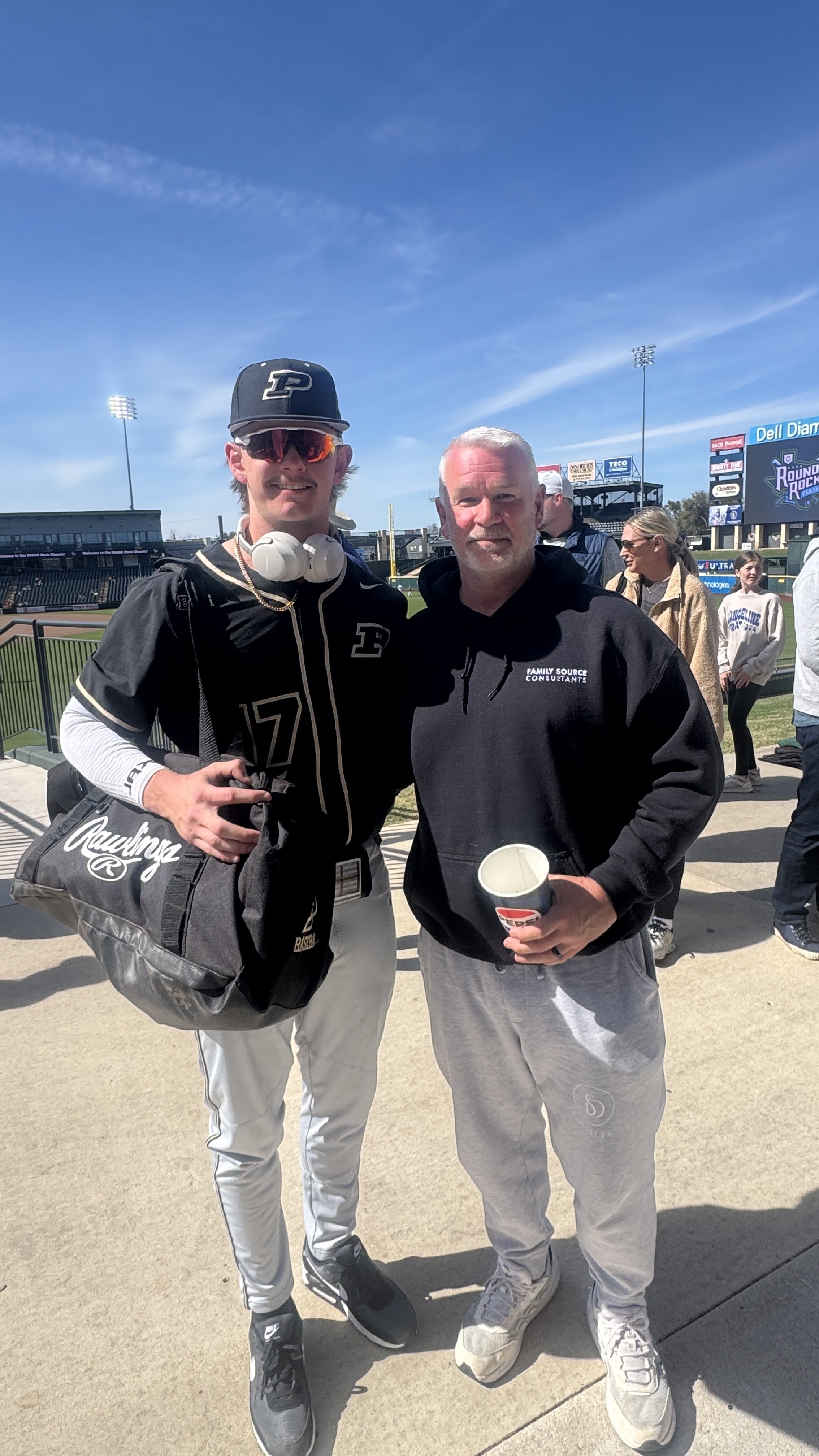 A young man in a baseball uniform stands on a sports field with an older man in casual clothing. The young man is wearing sunglasses, a black cap with a 'P' logo, and a black jersey with the number 17. They are posing for a photo, and there are other
