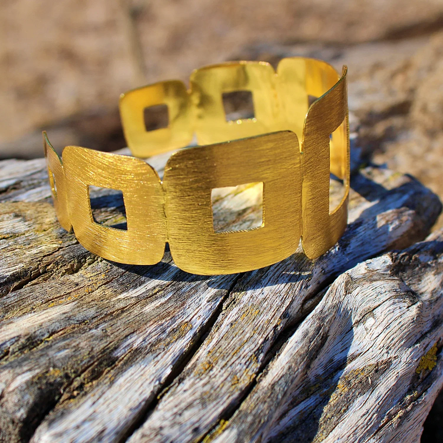 Gold-colored bracelet with square cutouts resting on weathered wooden surface outdoors.