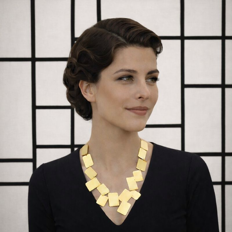 Woman with short dark hair wearing a black top and a large gold geometric necklace, standing in front of a black and white grid-patterned background.