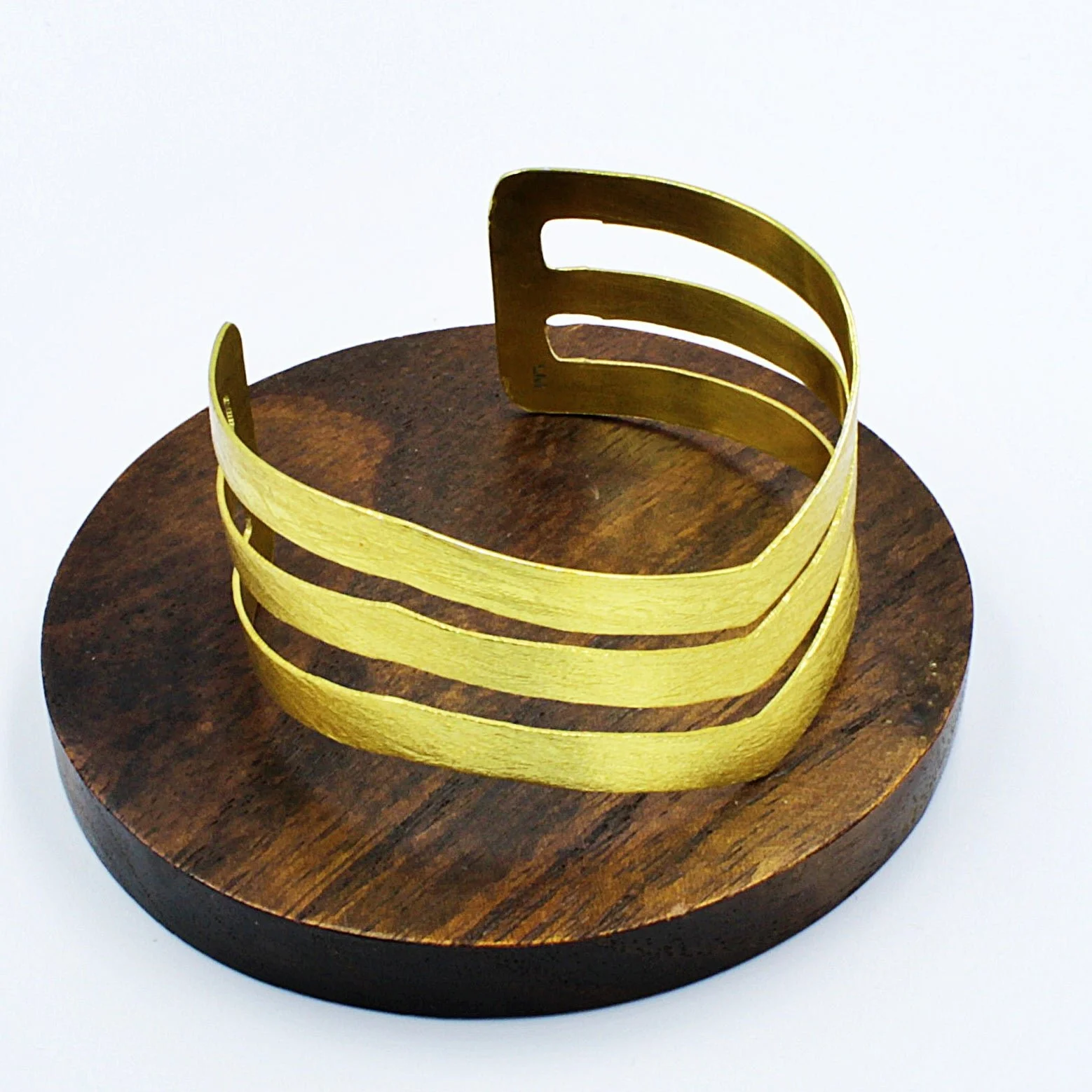 Gold metallic cuff bracelet with layered open design on a round wooden display stand.