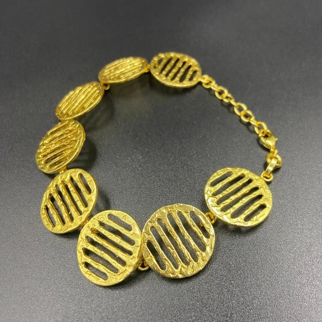 Gold statement bracelet with round, textured and striped oval links, laid on a dark surface.
