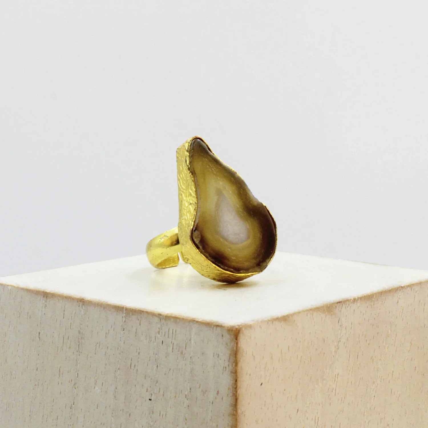 A gold ring featuring a large, teardrop-shaped, semi-precious stone with brown, beige, and white swirling patterns, displayed on a white pedestal against a plain background.