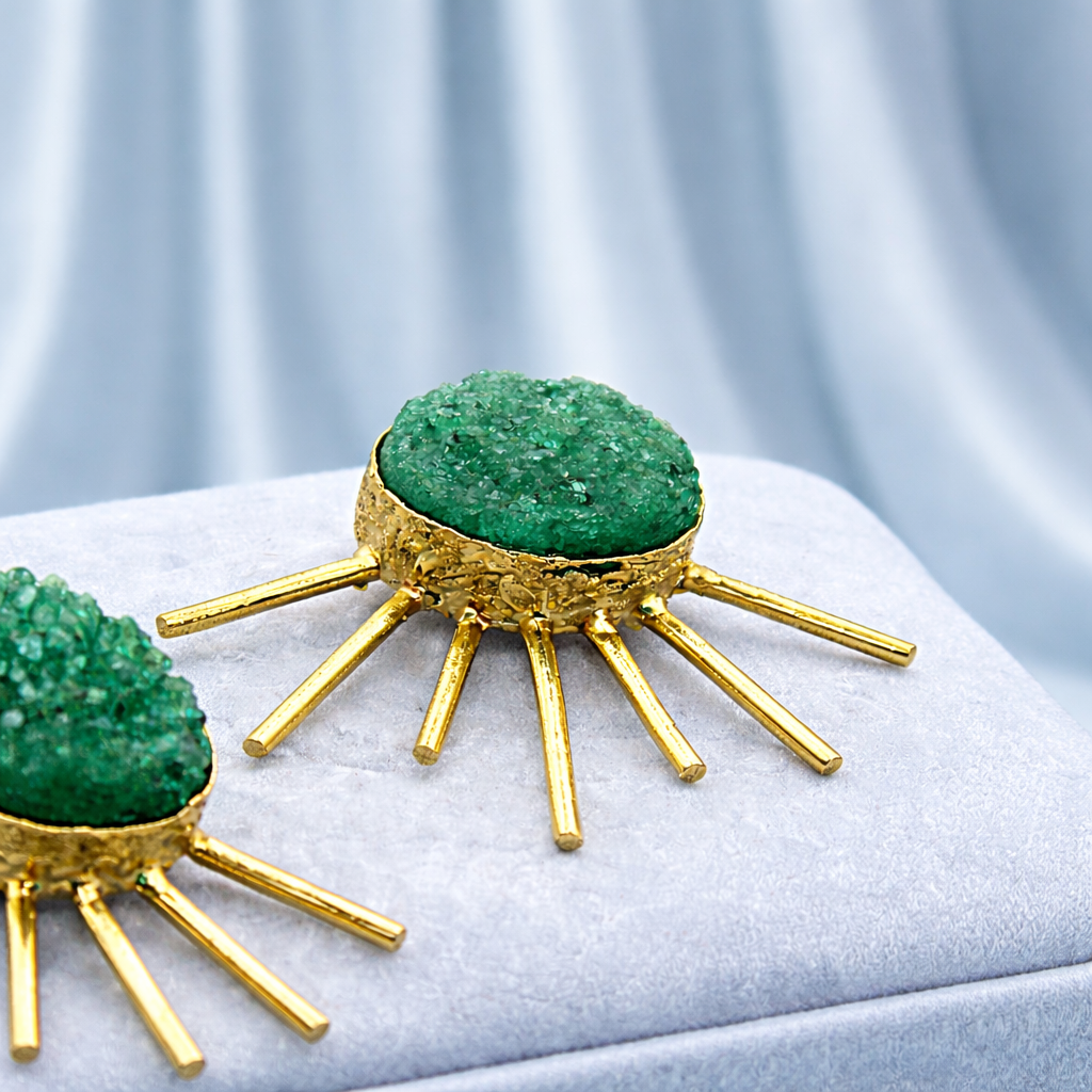Close-up of two gold jewelry pieces with green stone embellishments on white velvet fabric, set against a light gray background.