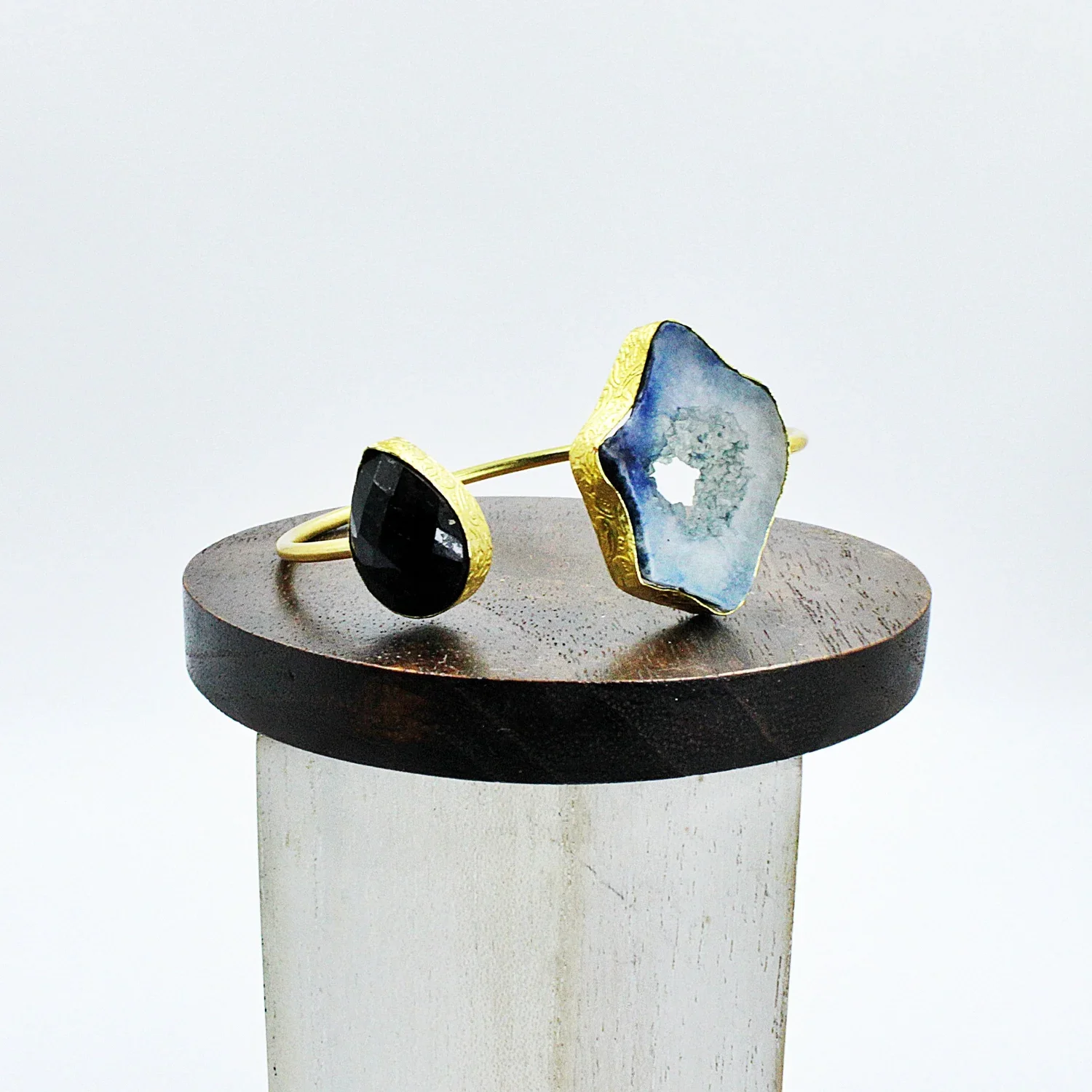 Gold bracelet with two large geode stones, one black and one blue, displayed on a wooden stand.