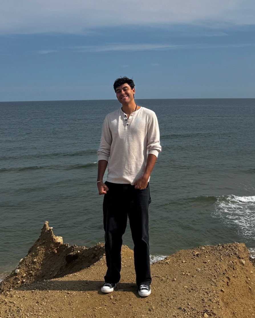 Young man standing on a rocky formation by the ocean, smiling, with water and sky in the background.