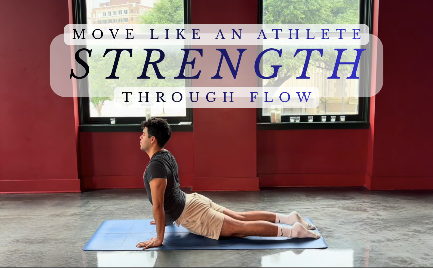 A man in a black t-shirt, beige shorts, and white socks practicing yoga in Child's Pose on a blue mat inside a room with red walls and large windows showing trees outside. Overlay text reads "Move like an athlete, strength through flow."