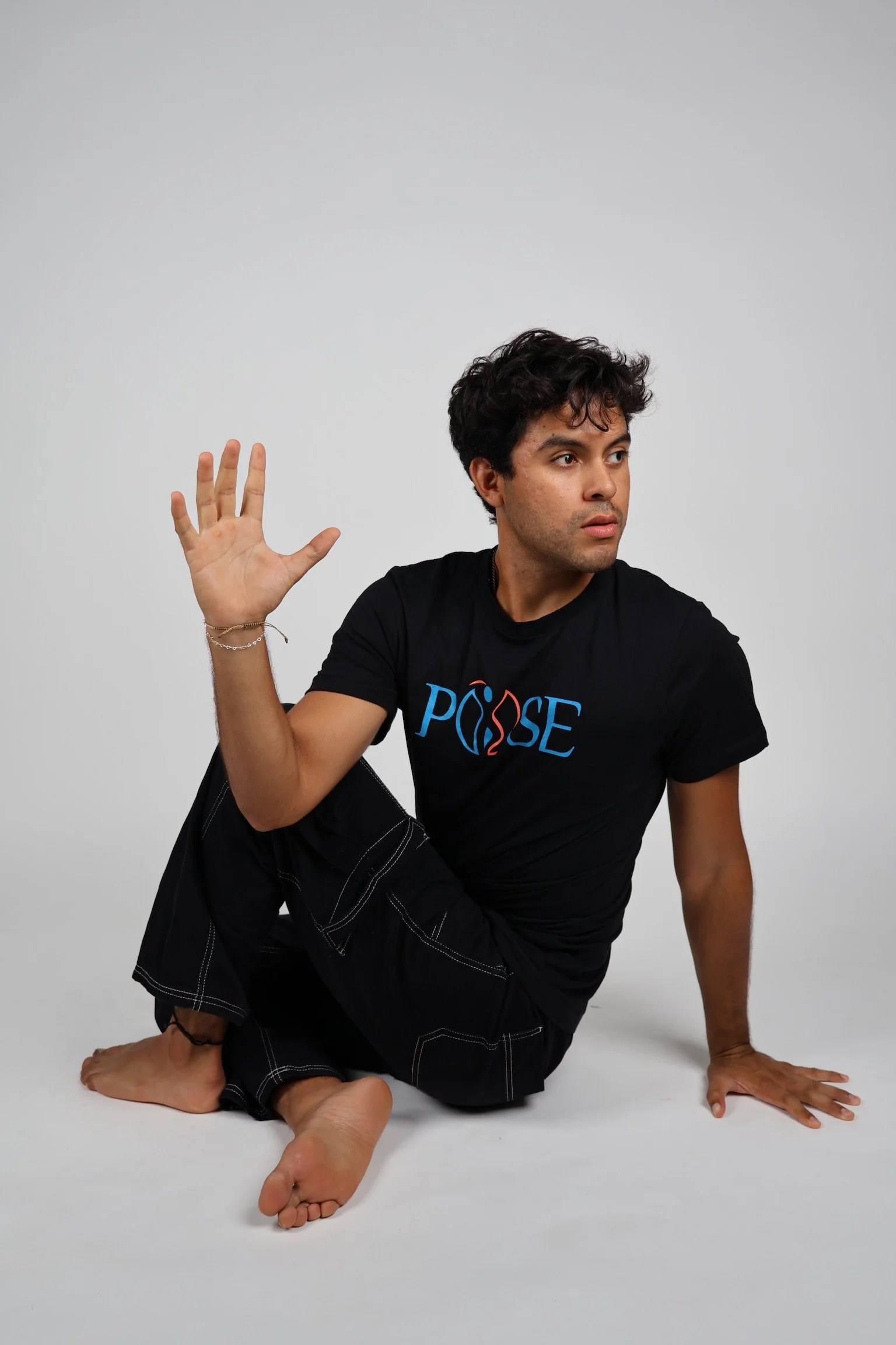A young man with dark hair and expressive eyes performing a flexible yoga pose on a plain white background. He is kneeling on one knee with the other leg bent, and one arm is raised with the hand open and fingers spread, while the other arm touches the ground for support. He is wearing a black T-shirt with the word "POSSE" in colorful letters and black pants with white stitching.