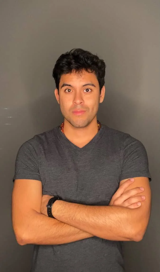 Young man with dark hair and brown eyes, wearing a dark gray t-shirt, crossing his arms, standing against a plain gray background.