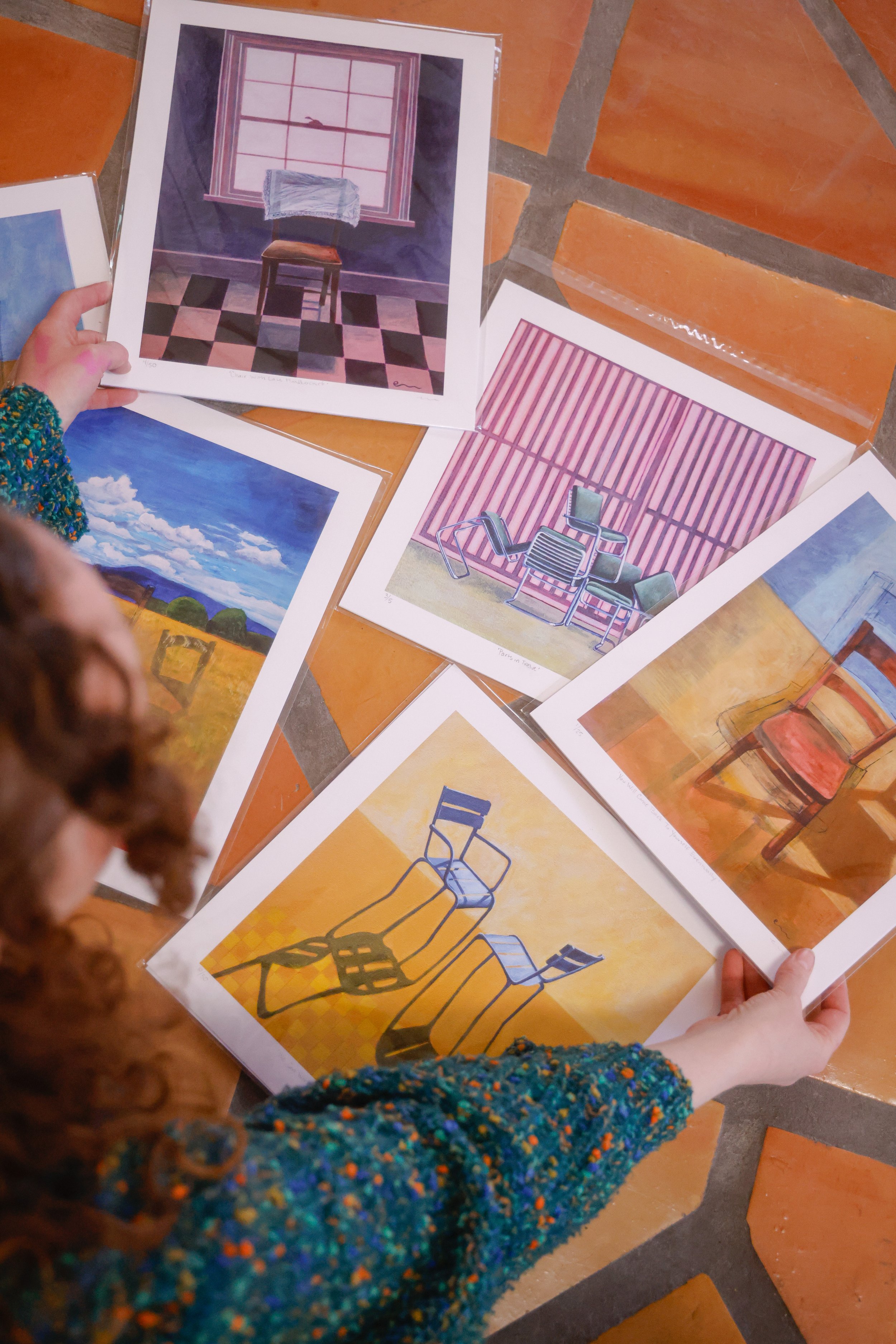 Varias láminas con pinturas de muebles y paisajes, algunas sostenidas por una persona de cabello rizado con un suéter multicolor, en un suelo de baldosas de color terracota.