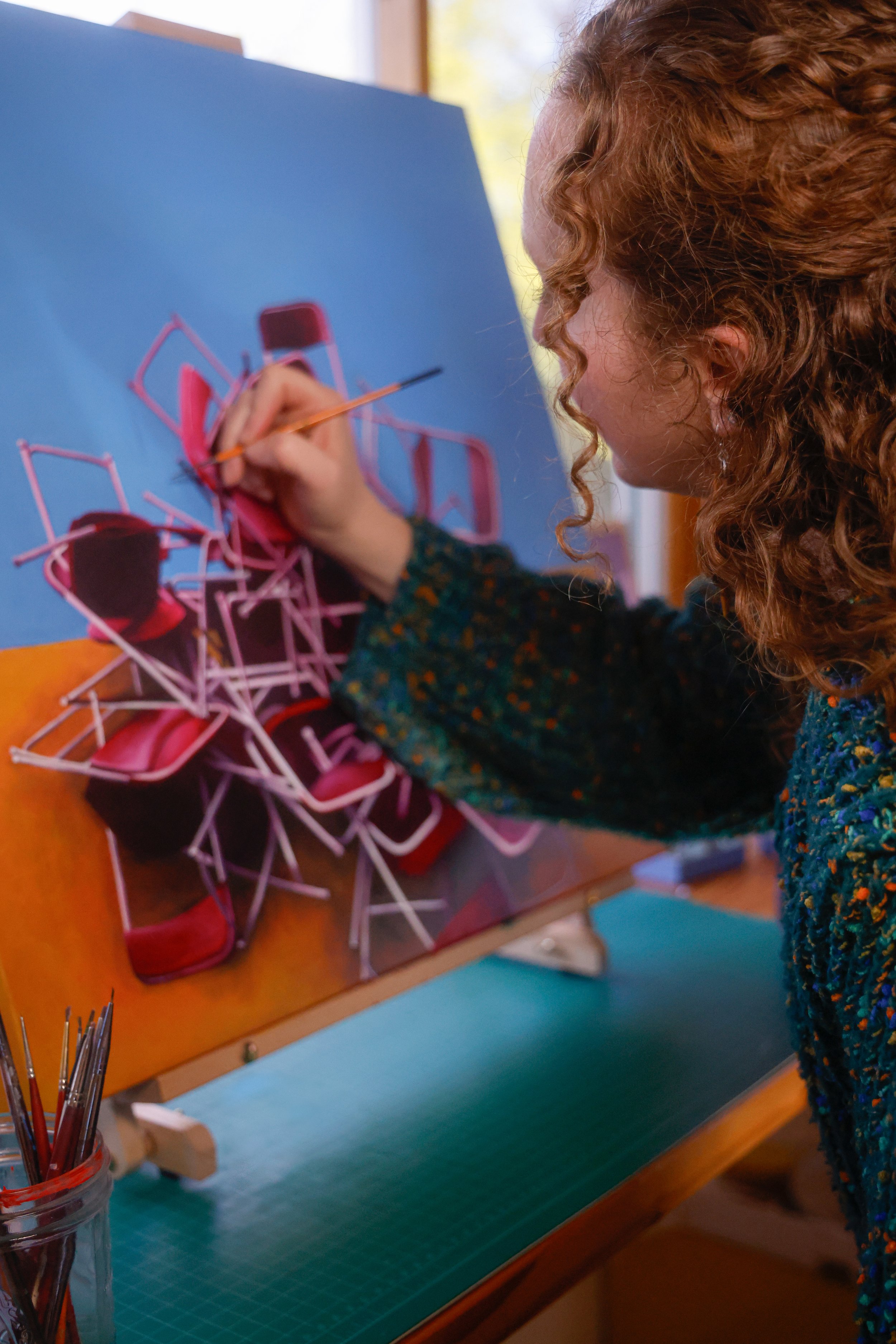 Una mujer con cabello rizado pintando una escultura de sillas con un pincel en un caballete en un espacio con luz natural.